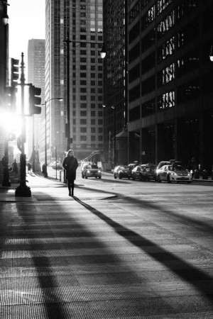Black and white image of a person walking down a city street with tall buildings and strong shadows.